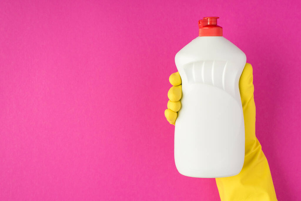 Photo de la main en gant de caoutchouc jaune tenant la bouteille de gel détergent sans étiquette sur fond rose isolé avec espace vide - Photo, image