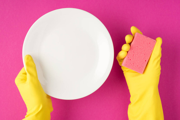 Πάνω όψη φωτογραφία των χεριών σε κίτρινα λαστιχένια γάντια που κρατούν λευκό καθαρό πιάτο και ροζ επίθεμα καθαρισμού σε απομονωμένο ροζ φόντο με κενό χώρο - Φωτογραφία, εικόνα