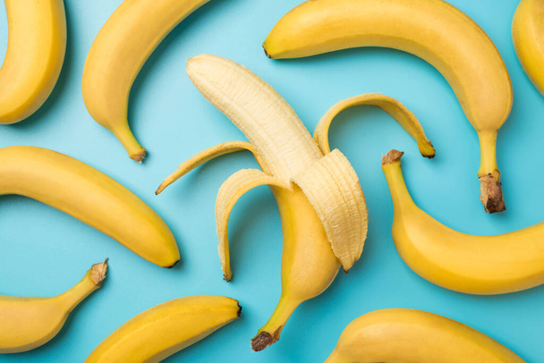 Top view photo of one peeled banana in the middle and scattered unpeeled bananas on isolated pastel blue background - Foto, afbeelding
