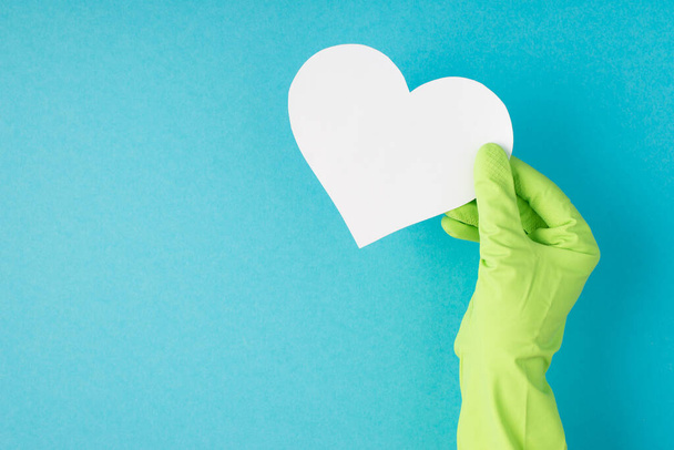 Фотографія рук зверху в зеленій гумовій рукавичці, що тримає біле паперове серце на ізольованому пастельно-блакитному тлі з порожнім простором
 - Фото, зображення