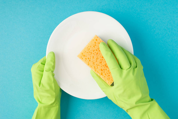 Foto de vista superior de las manos en guantes de goma verde que sostienen la almohadilla de fregar amarilla y la placa blanca limpia sobre fondo azul pastel aislado - Foto, imagen