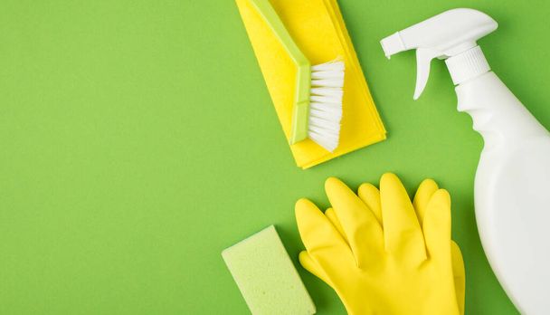 Top view photo of white spray detergent bottle without label sponge brush on folded yellow napkin and yellow rubber gloves on isolated green background with copyspace - Photo, Image