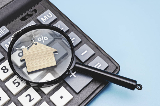 The concept of mortgage, sale and rental of housing and real estate. Mortgage credit lending. Calculator with a wooden house under a magnifying glass on a blue background. Copy space - Photo, Image