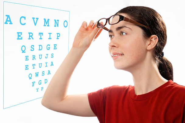 Ophtalmologie. Portrait d'une jeune belle femme enlevant ses lunettes et regardant un diagramme pour vérifier sa vision. Le concept des yeux sains et le test optique. - Photo, image