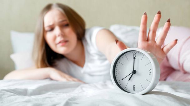 Overslept young woman awake in pajamas turns off alarm clock in hurry. Morning routine and wake up late from alarm clock in bed. Woman is late in panic after Unhealthy sleep. Long web banner - 写真・画像