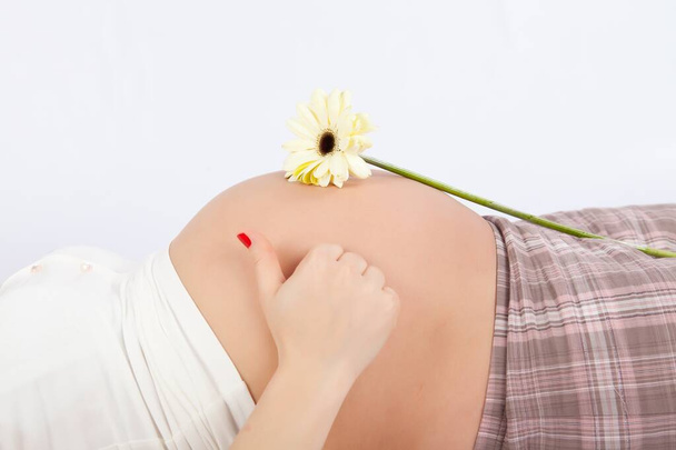 беременная женщина лежит на кровати на белом фоне и показывает большие пальцы вверх. желтый цветок на животе - Фото, изображение