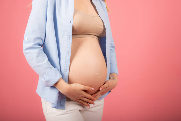 Beautiful pregnant woman in a blue shirt stroking her belly on a pink background. - Photo, Image