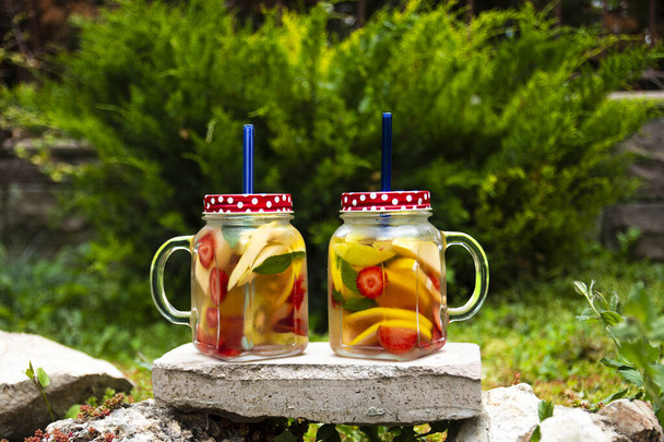 Две стеклянные банки, вкусный детоксикационный напиток с красными, оранжевыми и желтыми фруктами, стоящими на камне. Селективные фокус-очки. - Фото, изображение