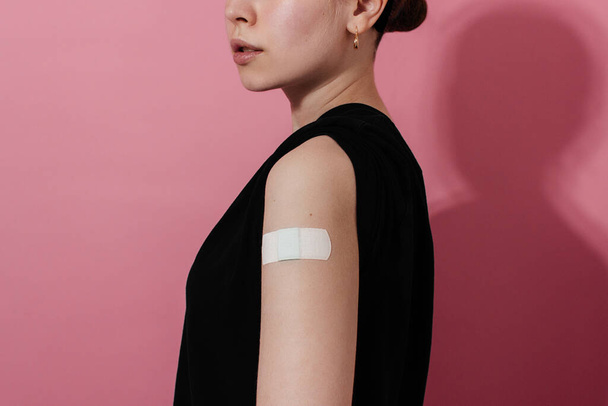 Улыбающаяся молодая женщина в футболке с гипсом на плече после коронавирусной вакцины, розовый фон, копия пространства. Вакцинация, иммунизация, концепция профилактики гриппа. Получение вакцины Ковид-19 - Фото, изображение