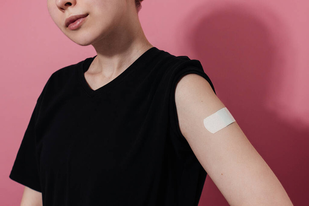 Smiling young woman in t-shirt showing plaster on shoulder after coronavirus vaccine, pink background. Vaccination, immunization, flu prevention concept. Getting Covid-19 vaccine - 写真・画像