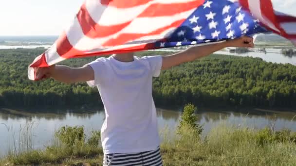 Blonde boy waving national USA flag outdoors over blue sky at the river bank - 映像、動画