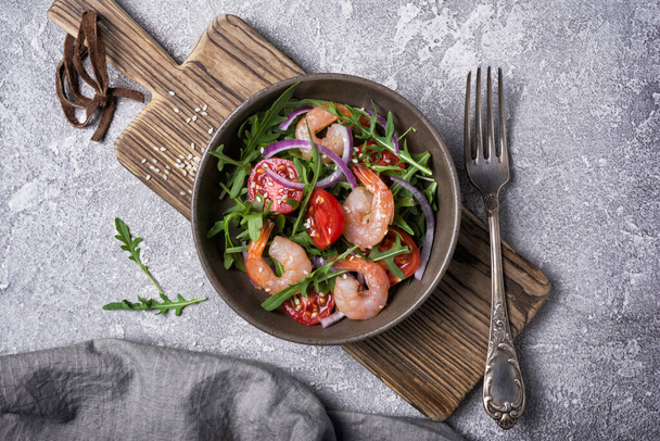 Top view of healthy and tasty food. Salad bowl with fresh arugula leaves, cherry tomatoes, shrimps, onion and sesame on grey concrete background - 写真・画像