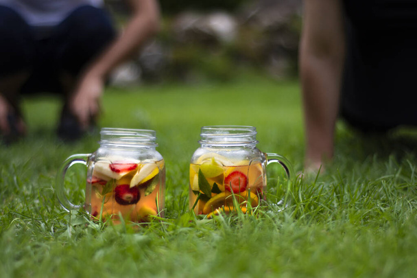 Δύο Jars από γυαλί, νόστιμο ρόφημα αποτοξίνωσης με κόκκινα, πορτοκαλί και κίτρινα φρούτα, στέκεται στο γρασίδι. Επιλεκτικά γυαλιά εστίασης. - Φωτογραφία, εικόνα
