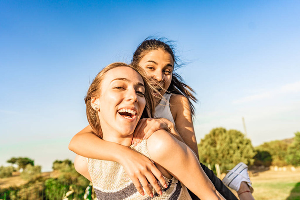 Two girls best friends having fun outdoor in a green nature park looking at camera laughing and joking. Young brunette woman jumping on back of her blonde girlfriend surprising her. Happy students - Foto, imagen