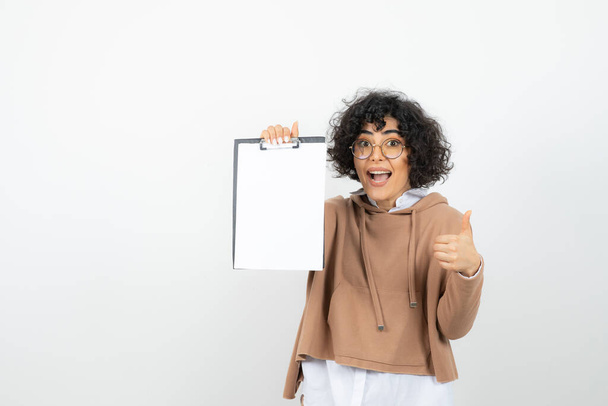Portrait of young woman in glasses with empty clipboard giving thumbs up. High quality photo - Photo, Image