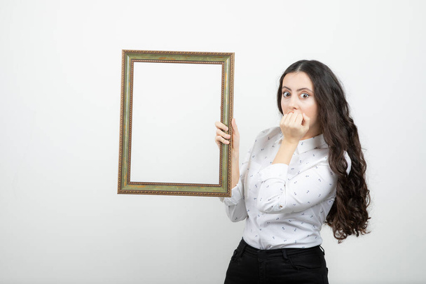 Mujer joven de pie y sosteniendo el marco de la imagen contra la pared blanca. Foto de alta calidad - Foto, Imagen