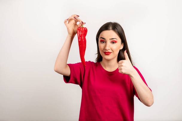 Μελαχρινή γυναίκα κρατάει κόκκινη καυτερή πιπεριά τσίλι και δείχνει τον αντίχειρα ψηλά. Υψηλής ποιότητας φωτογραφία - Φωτογραφία, εικόνα