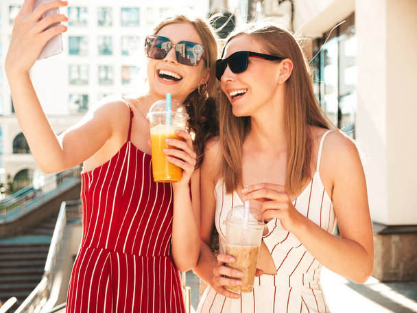 Két fiatal, gyönyörű, mosolygós, divatos nyári ruhás hipster nő. Gondtalan nők pózolnak a szabadban Pozitív modellek ivás friss koktél smoothie ital műanyag pohár szalmával.Készít szelfi - Fotó, kép