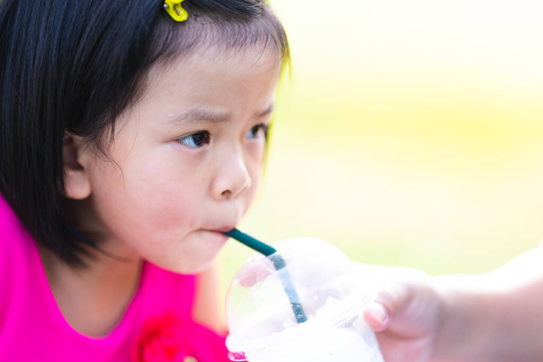 甘いココナッツスムージーを吸うアジアの子供の女の子の頭のショット.母親は娘にグラスを持ってる。赤ちゃんはチューブで冷たい水を飲む。夏時間だ暑い日だ。コピースペース。4 〜 5歳の子供たち. - 写真・画像