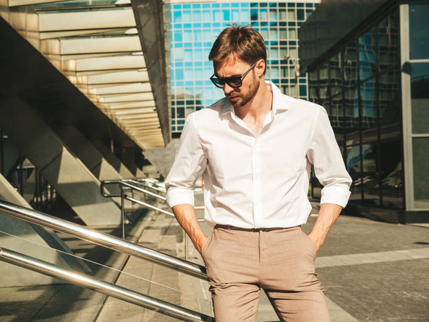 Portrait of handsome confident stylish hipster lambersexual model.Modern man dressed in white shirt. Fashion male posing in the street background near skyscrapers in sunglasses. Outdoors at sunset  - Fotó, kép