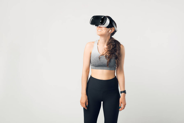 Photo of young sport woman wearing sportswear and VIRTUAL REALITY HELMET over white background. - Foto, Imagen