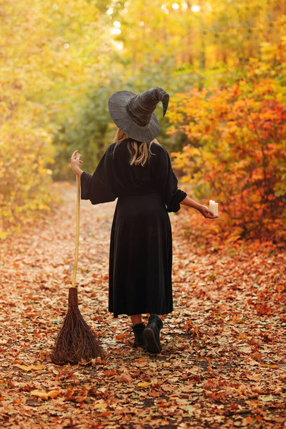 Visszapillantás egy névtelen boszorkányjelmezes nőre seprűvel az ösvényen, őszi levelekkel borítva Halloweenkor az erdőben. - Fotó, kép