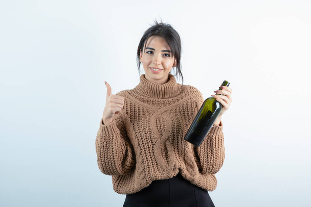 Πορτρέτο της νεαρής γυναίκας κρατώντας ένα μπουκάλι κρασί και δίνοντας αντίχειρες επάνω σε λευκό φόντο. Υψηλής ποιότητας φωτογραφία - Φωτογραφία, εικόνα