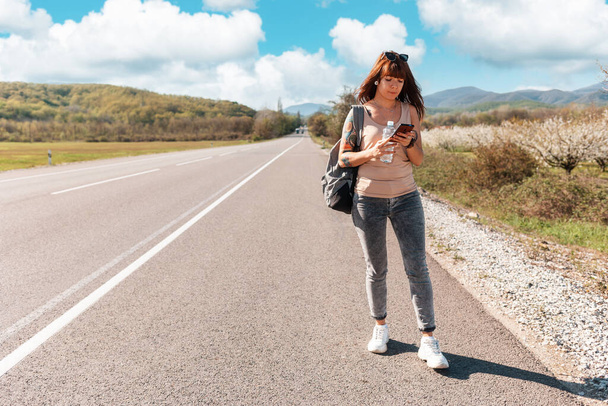 Valkoihoinen nuori nainen, jolla on reppu ja tatuoitu käsi seisoo tyhjällä tiellä ja käyttää älypuhelinta. Kopioi tilaa. Käsite paikallisen matkustamisen ja internet kartta sovellus. - Valokuva, kuva