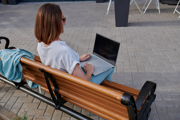 κοκκινομάλλα γυναίκα κάθεται στον πάγκο και εργάζεται σε φορητό υπολογιστή - Φωτογραφία, εικόνα