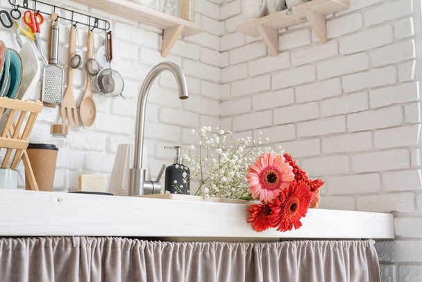 Rustykalne wnętrze kuchni z białą ceglaną ścianą i białymi drewnianymi półkami. Czerwony i różowy gerbera stokrotki w zlewozmywaku - Zdjęcie, obraz