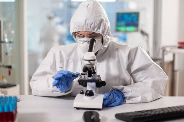 Ученый в регулировке костюмов ppe и просматривает лабораторный микроскоп - Фото, изображение