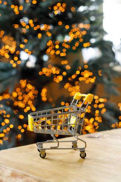New Year and Christmas background. Small grocery cart stands on wooden table against background of bokeh garlands on christmas tree - Φωτογραφία, εικόνα