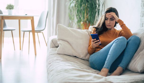 Frustrated and depressed young brunette woman is crying with a smartphone in hands while she sitting on the couch at home - Foto, afbeelding