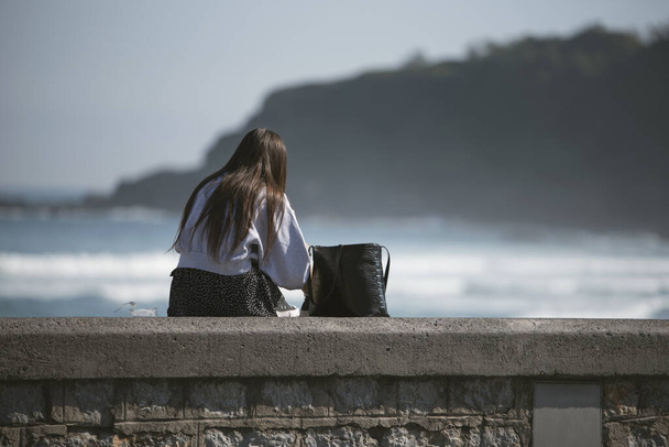 Вид сзади молодой брюнетки, сидящей на каменном заборе в солнечный день - Фото, изображение