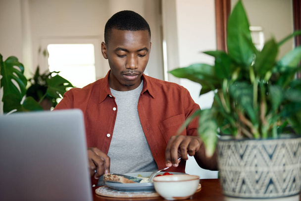 彼のダイニングルームのテーブルに座って、朝食を食べながらノートパソコンで何かを見ている若いアフリカ人男性 - 写真・画像