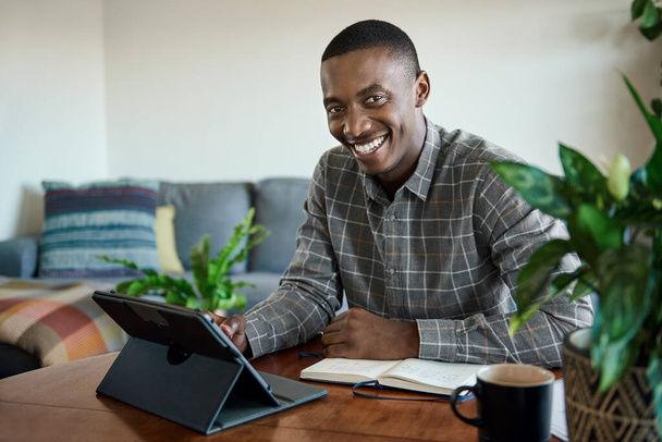 Portrait of a smiling young African businessman working remotely from home at a living room table using a digital tablet - Fotoğraf, Görsel