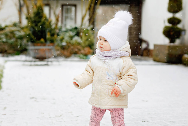 Счастливая маленькая девочка делает первые шаги на улице зимой через снег. Милый малыш учится ходить. Ребенок веселится в холодный снежный день. Первый снег, активность. Зимняя прогулка на природе - Фото, изображение