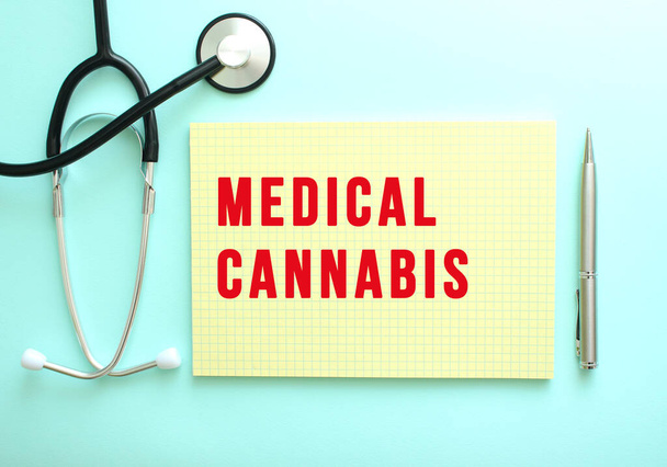 The red text MEDICAL CANNABIS is written in a yellow pad that lies next to the stethoscope on a blue background. Medical concept - Photo, image