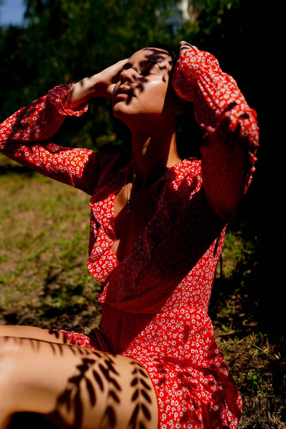 Νεαρή γυναίκα έξω. Φως και σκιά. νεαρό κορίτσι με κόκκινο φόρεμα καλυμμένο με ασυνήθιστη σκιά από φύλλα - Φωτογραφία, εικόνα