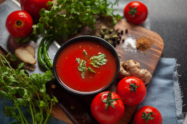 Hint ev yapımı taze ve sağlıklı domates çorbası taze kişniş yaprakları, malzemeler ve otlarla süslenmiş, ahşap arka planda siyah bir kasede servis ediliyor.. - Fotoğraf, Görsel