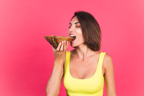 Beautiful fit woman in yellow bright fitting sportswear on pink background happy hold healthy avocado sandwich - Photo, Image