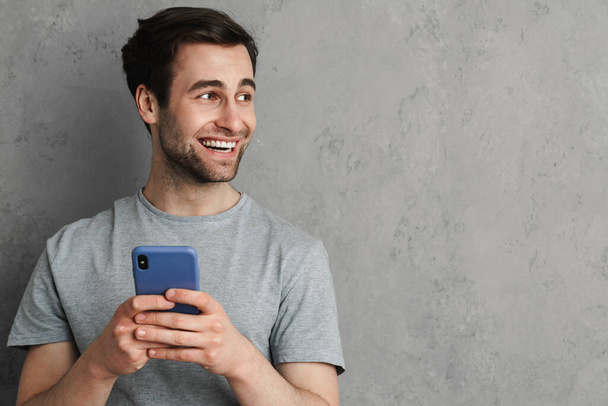 Retrato de um jovem sorridente segurando telefone celular e olhando para o espaço de cópia isolado sobre fundo cinza - Foto, Imagem