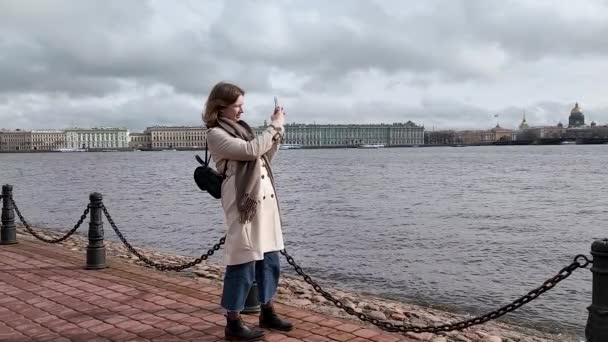 Uma jovem mulher com um telefone celular em suas mãos tira fotos do dique do rio Neva em São Petersburgo, Praça do Palácio e Catedral de São Isaacs. - Filmagem, Vídeo
