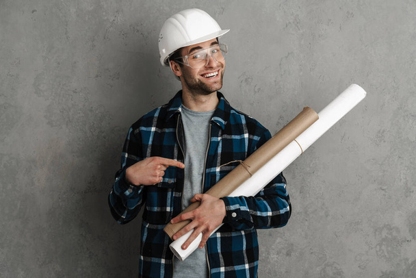 Joyful builder man in helmet and goggles holding and pointing finger at drawings isolated over grey wall - Foto, Bild