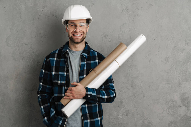 Young handsome civil engineer in hardhat holding blueprints standing isolated over gray background - Fotoğraf, Görsel