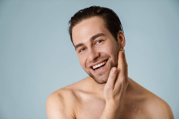 Close up of a smiling brunette shirtless man with stubble over blue background, touching face - Fotó, kép