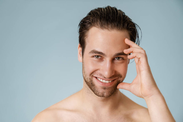 Close up of a smiling brunette shirtless man with stubble over blue background, touching face - Foto, Imagen