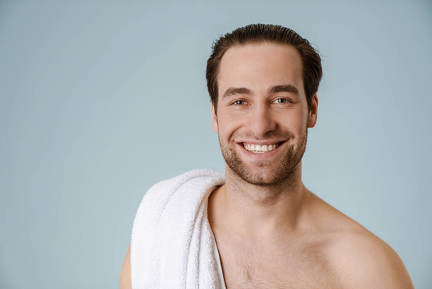 Shirtless white man smiling at camera while posing with towel isolated over blue wall - Fotó, kép