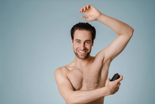 Handsome smiling nude man spraying deodorant isolated over blue background - Foto, afbeelding