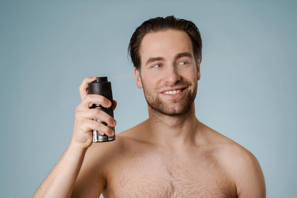 Handsome smiling nude man spraying deodorant isolated over blue background - Foto, Imagen